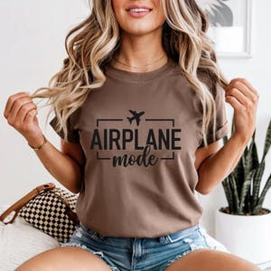 May include: A brown t-shirt with the words "AIRPLANE mode" in black, with a small airplane graphic above the text. The shirt is short-sleeved and appears to be made of a soft material.