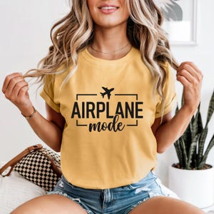 May include: Mustard yellow t-shirt with the words "AIRPLANE mode" in black, along with a small airplane graphic. The text is enclosed in a rectangular frame. The shirt has short sleeves and a crew neck.