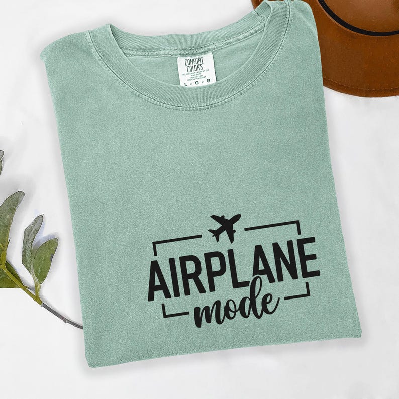 May include: A light green t-shirt with the words "AIRPLANE mode" in black, along with a small airplane graphic. The shirt is folded and has a tag that says "COMFORT COLORS".