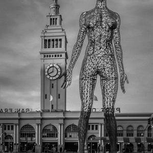 Puede incluir: Una gran escultura en blanco y negro de una figura humana construida con una malla de alambre densa e intrincada. La figura se alza sobre una base circular frente a un edificio con torre de reloj de varios niveles y arcos.