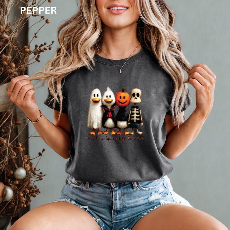 K&ouml;nnte beinhalten: Dunkelgraues T-Shirt mit einem Halloween-Motiv von vier Cartoon-Enten, die als Geist, Pinguin, K&uuml;rbis und Skelett verkleidet sind. Der Text "Just a Spooky Goose" ist darunter gedruckt. Das Wort "PEPPER" ist oben links aufgedruckt.