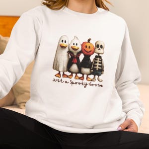 K&ouml;nnte beinhalten: Wei&szlig;es Sweatshirt mit einem Halloween-Motiv. Die Grafik zeigt vier Cartoon-Figuren: ein Geist, ein Pinguin, ein K&uuml;rbis und ein Skelett. Der Text "Just a Spooky Goose" ist darunter gedruckt. Das Sweatshirt hat lange &Auml;rmel.