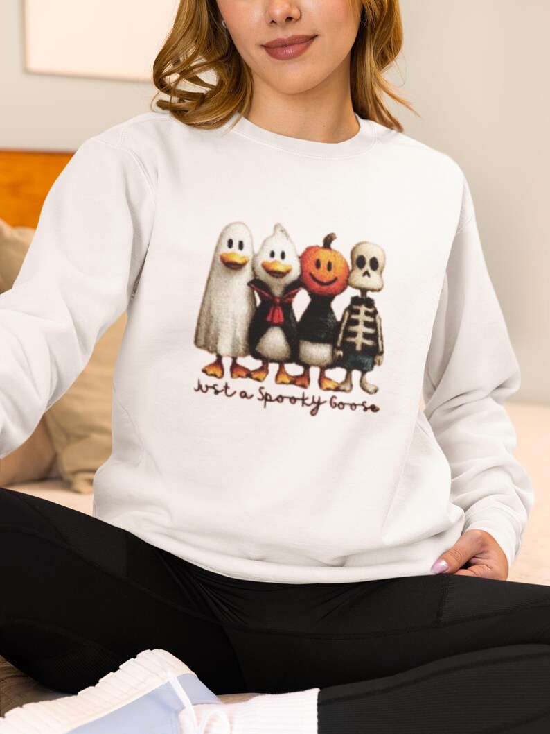 K&ouml;nnte beinhalten: Wei&szlig;es Sweatshirt mit einem Halloween-Motiv. Die Grafik zeigt vier Cartoon-Figuren: ein Geist, ein Pinguin, ein K&uuml;rbis und ein Skelett. Der Text "Just a Spooky Goose" ist darunter gedruckt. Das Sweatshirt hat lange &Auml;rmel.