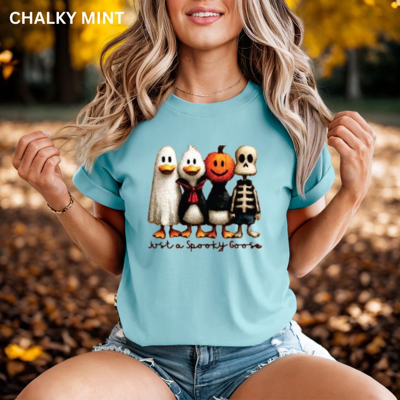 K&ouml;nnte beinhalten: Hellblaues T-Shirt mit einem Halloween-Motiv. Das Design zeigt vier Cartoon-G&auml;nse, die als Geist, Pinguin, K&uuml;rbis und Skelett verkleidet sind. Der Text "Just a Spooky Goose" ist darunter gedruckt.