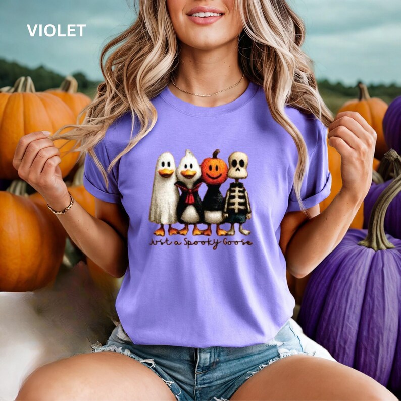 K&ouml;nnte beinhalten: Violettes T-Shirt mit einem Halloween-Motiv, das einen Geist, eine Ente, einen K&uuml;rbis und ein Skelett zeigt. Der Text "Just a Spooky Goose" steht darunter. Im Hintergrund sind K&uuml;rbisse zu sehen.