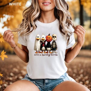 K&ouml;nnte beinhalten: Wei&szlig;es T-Shirt mit einem Halloween-Motiv. Das Design zeigt einen Geist, einen Pinguin, einen K&uuml;rbis und ein Skelett, mit dem Text "Just a Spooky Goose" darunter. Das T-Shirt ist aus weichem Material.