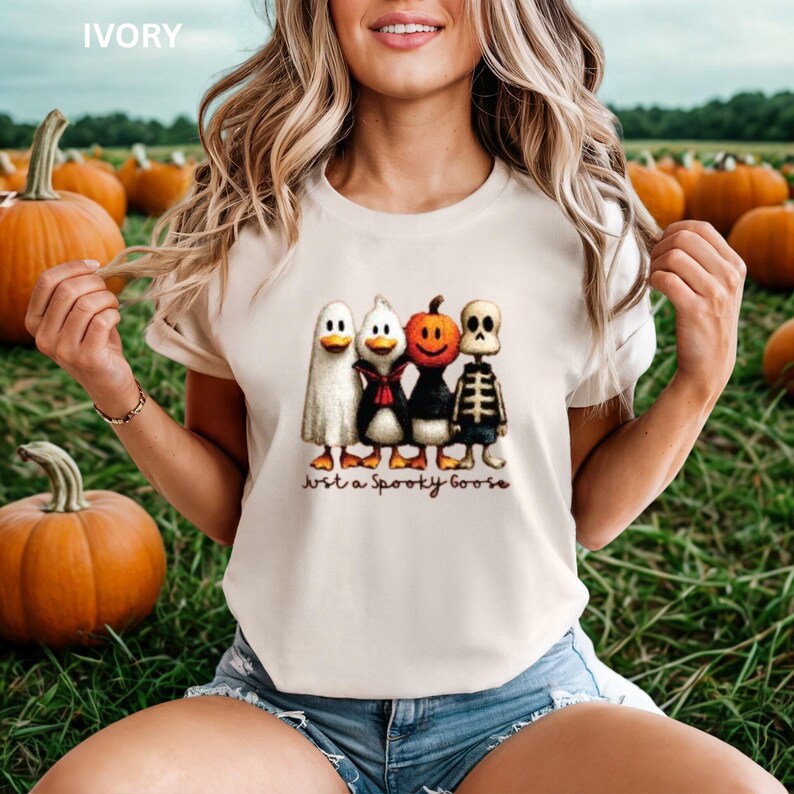 K&ouml;nnte beinhalten: Elfenbeinfarbenes T-Shirt mit einem Halloween-Motiv von vier Cartoon-Charakteren: einem Geist, einer Ente, einem K&uuml;rbis und einem Skelett. Der Text "Just a Spooky Goose" ist darunter gedruckt. Der Hintergrund zeigt K&uuml;rbisse.