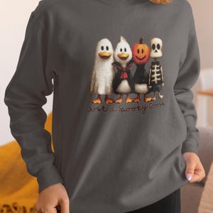 K&ouml;nnte beinhalten: Dunkelgraues Sweatshirt mit einem Halloween-Motiv. Die Grafik zeigt vier Enten in gruseligen Kost&uuml;men: ein Geist, ein Vampir, ein K&uuml;rbis und ein Skelett. Der Text "Just a Spooky Goose" steht darunter.