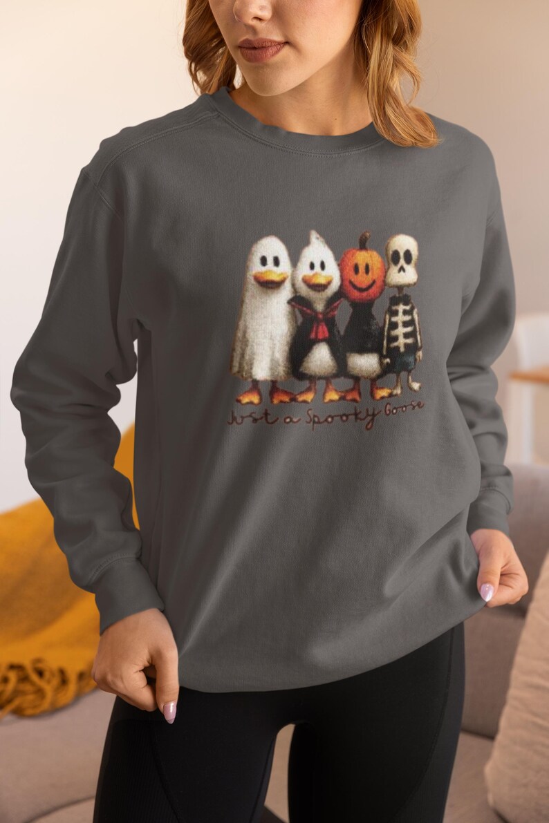 K&ouml;nnte beinhalten: Dunkelgraues Sweatshirt mit einem Halloween-Motiv. Die Grafik zeigt vier Enten in gruseligen Kost&uuml;men: ein Geist, ein Vampir, ein K&uuml;rbis und ein Skelett. Der Text "Just a Spooky Goose" steht darunter.
