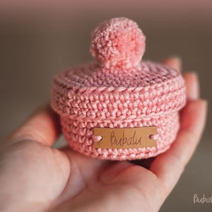 May include: A handmade pink crochet box with a matching pom-pom on the lid. The box has a small wooden tag that reads "Bubalu". The image also includes the text "Porta carinho" in a script font.