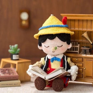 May include: A crocheted Pinocchio doll wearing a yellow hat with a blue band and a red feather. The doll has black hair, a red vest, and is reading a book. The doll is sitting in front of a wooden cabinet and a small table with books.