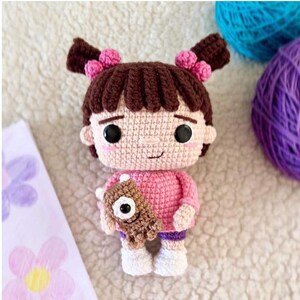 K&ouml;nnte beinhalten: Handgefertigte H&auml;kelpuppe, die einer Zeichentrickfigur &auml;hnelt, mit einem rosa Pullover, lila Shorts und wei&szlig;en Schuhen. Die Puppe hat braune Haare, die mit rosa Akzenten zu Z&ouml;pfen geflochten sind, und h&auml;lt ein braunes Monster. Wollkn&auml;uel in T&uuml;rkis und Lila sind im Hintergrund.