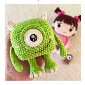 K&ouml;nnte beinhalten: Geh&auml;keltes gr&uuml;nes Monster mit einem gro&szlig;en Auge, zwei H&ouml;rnern und kleinen Armen und Beinen. Im Hintergrund befindet sich eine geh&auml;kelte Puppe eines M&auml;dchens mit braunen Haaren und rosa Schleifen. Das Monster hat ein l&auml;chelndes Gesicht mit rosa Wangen. Handgemachtes Spielzeug.