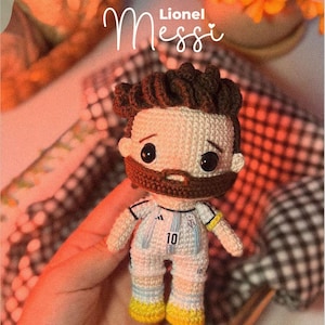 Puede incluir: Muñeco de ganchillo hecho a mano de Lionel Messi, con un uniforme de fútbol blanco y celeste con el número 10. El muñeco tiene pelo castaño y barba. El nombre "Lionel Messi" está escrito encima.