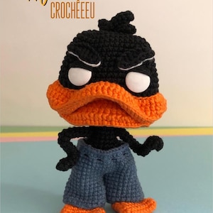 May include: A handmade crochet Daffy Duck figure. The figure has a black head with white eyes, an orange beak, and is wearing blue shorts and orange feet. The text "Daffy Duck CROCHEEEU" is in the upper left corner.