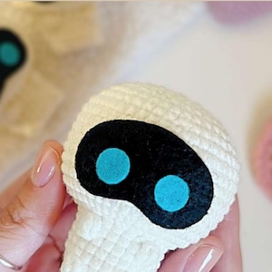 Puede incluir: Una cabeza de muñeca de ganchillo blanca con una cara ovalada negra y dos círculos azul turquesa. La muñeca está hecha de un hilo suave y texturizado. El fondo incluye un cuaderno de espiral y un corazón de ganchillo rosa.