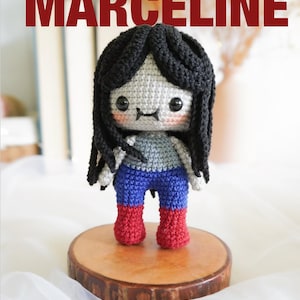 Puede incluir: Muñeca amigurumi hecha a mano llamada Marceline, con cabello de hilo negro, una parte superior gris y azul, pantalones azules y botas rojas. La muñeca está tejida a ganchillo y se encuentra sobre una rodaja de madera. Las palabras "AMIGURUMI" y "MARCELINE" están escritas en rojo.
