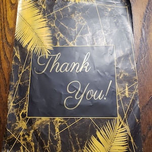Könnte beinhalten: Schwarzer und goldener Poly-Versandbeutel mit Marmormuster und goldenen Palmenblättern. Der Beutel hat ein schwarzes Quadrat mit dem Text "Thank You!" in goldenen Buchstaben.