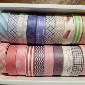 May include: A white box filled with rolls of decorative washi tape. The tape features various patterns, including stripes, geometric shapes, and floral designs, in shades of blue, purple, pink, and green. The rolls are approximately 1.5 inches in diameter.