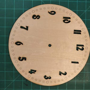 May include: A circular, unfinished wooden clock face featuring black numerals and small circular markings around the perimeter. Numbers 1 to 12 are present. A small hole is in the centre.