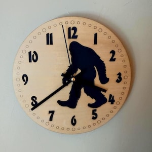 May include: A round wooden clock with a light wood grain finish. The clock face features black numerals and a black silhouette of Bigfoot. The clock hands are black, and the clock is mounted on a light gray wall.