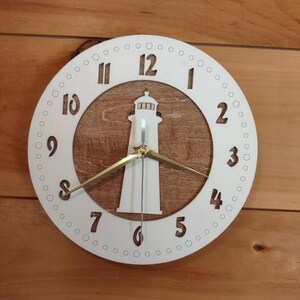 Puede incluir: Un reloj blanco con forma de faro con fondo de madera. La esfera del reloj presenta un anillo exterior blanco con números negros y un recorte de faro. Las manecillas del reloj son doradas.