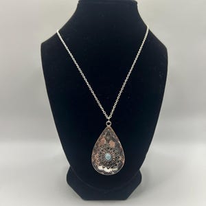 May include: Silver-toned pendant necklace with a teardrop-shaped pendant. The pendant features an intricate floral design with a small, light blue stone in the center. The necklace is displayed on a black mannequin.