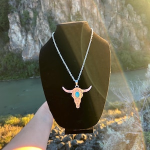 May include: A silver-toned metal necklace featuring a pendant shaped like a steer skull. The pendant is decorated with etched black line patterns and a central oval-shaped turquoise-colored stone. The pendant hangs from a thin silver-toned chain.