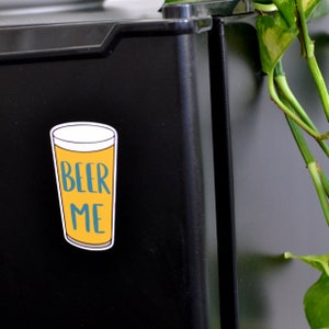 K&ouml;nnte beinhalten: Eine gelbe und blaue Cartoon-Illustration eines Bierglases mit dem Text "BEER ME" auf einem schwarzen K&uuml;hlschrank.