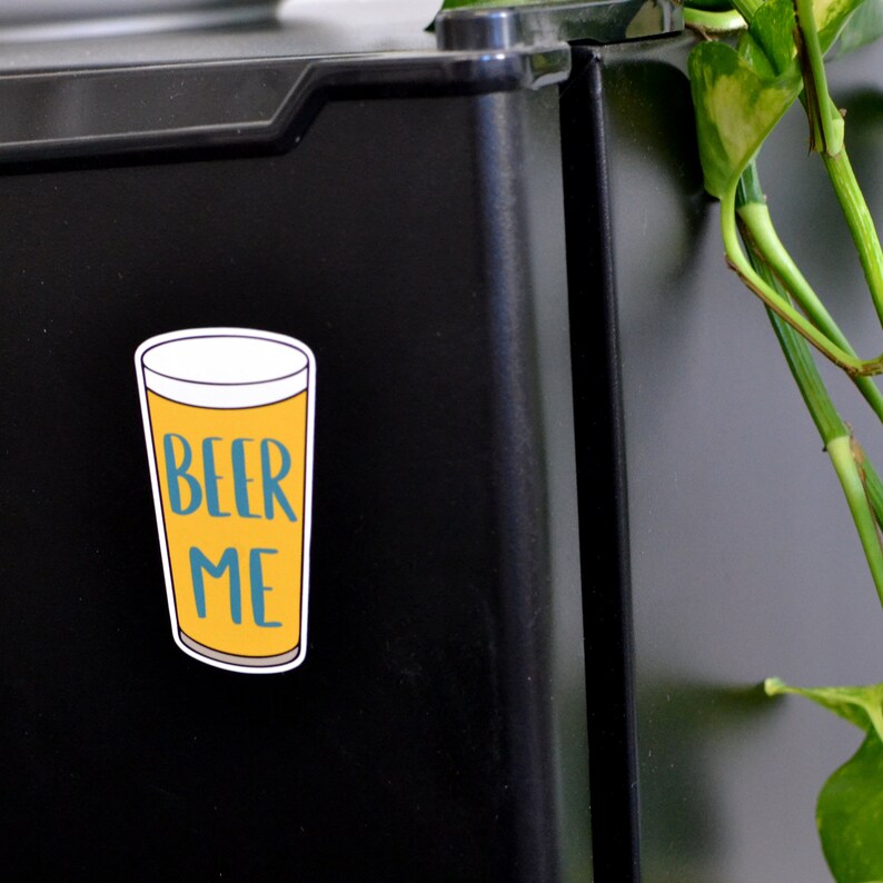 K&ouml;nnte beinhalten: Eine gelbe und blaue Cartoon-Illustration eines Bierglases mit dem Text "BEER ME" auf einem schwarzen K&uuml;hlschrank.