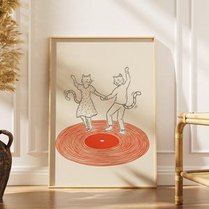 Puede incluir: Lámina artística enmarcada con una ilustración de dos gatos bailando sobre un disco de vinilo rojo. Los gatos están dibujados con líneas negras, tomados de la mano. El fondo es beige neutro, con marco dorado.
