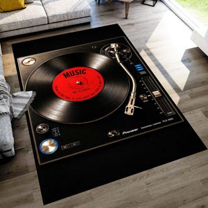 Op de afbeelding: Zwart rechthoekig vloerkleed met een gedetailleerde afbeelding van een vintage platenspeler. De plaat is rood met het woord "MUSIC" in het wit. Het vloerkleed heeft een gouden rand en de merknaam "Pioneer" is zichtbaar.