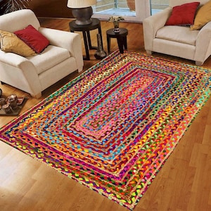 Peut inclure: Un tapis rectangulaire tressé multicolore avec un éventail de couleurs vives, notamment rose, rouge, jaune, vert et bleu. Le tapis présente un motif répétitif de sections tressées, créant une surface texturée. Le tapis est posé sur un sol en bois.