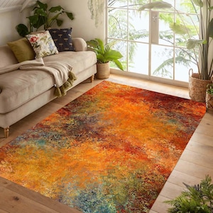 Op de afbeelding: Een rechthoekig vloerkleed met een abstract ontwerp. Het kleed heeft een mix van warme kleuren, waaronder oranje, rood, geel en vleugjes blauw en groen. Het ligt op een houten vloer in een woonkamer.