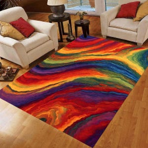 May include: A rectangular area rug with a vibrant, abstract design. The rug features a flowing pattern of layered colors, including red, orange, yellow, green, blue, and purple. The rug is placed on a hardwood floor, with two cream-colored armchairs visible.