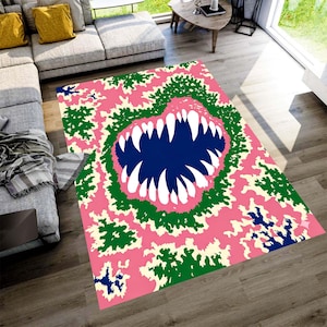 May include: A rectangular rug with a monster mouth design. The rug features a blue mouth with sharp white teeth, surrounded by green, pink, and cream-colored patterns. The rug is placed on a wooden floor.