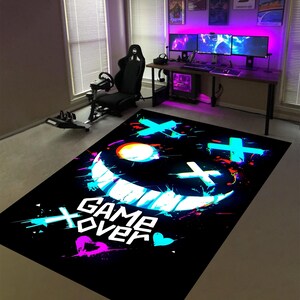 May include: Black rectangular rug with a neon-style smiley face design and the words "GAME OVER" in white. The rug features splashes of blue, pink, and orange, creating a vibrant, modern aesthetic. The rug is placed in a gaming room.