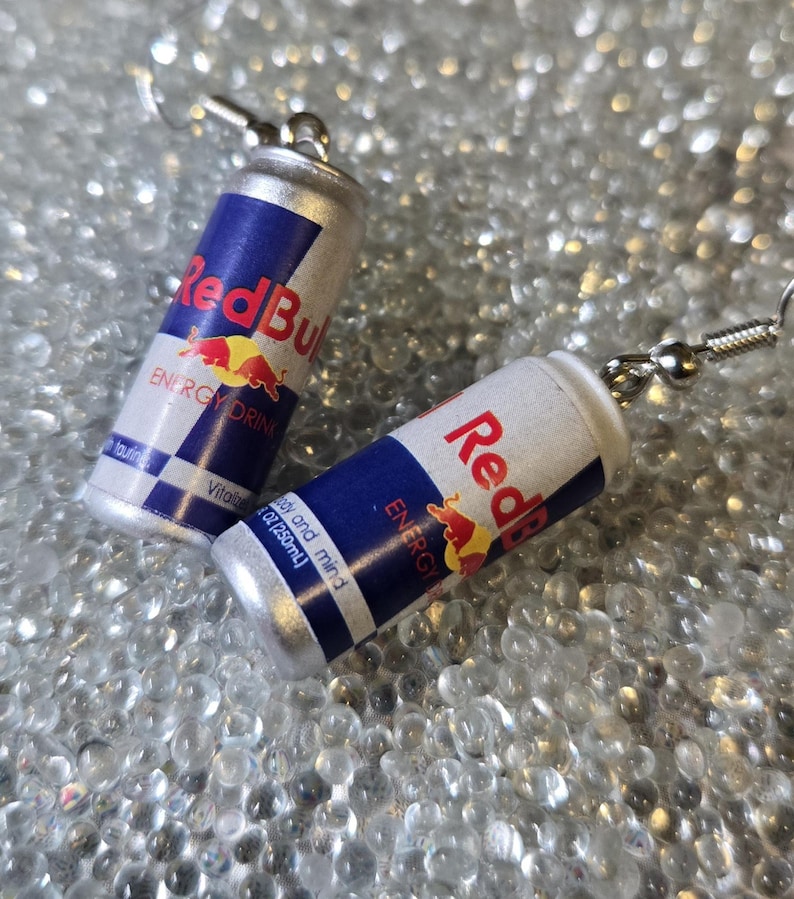 Puede incluir: Un par de pendientes novedosos con forma de mini latas de bebida energ&eacute;tica Red Bull. Las latas son plateadas, blancas y azules con el logotipo y el texto de Red Bull. Los pendientes tienen cierres de gancho de pez plateados.