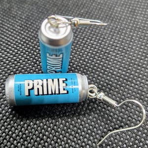 Puede incluir: Pendientes novedosos con forma de peque&ntilde;as latas de bebida energ&eacute;tica PRIME azules y plateadas. Los pendientes muestran la palabra "PRIME" en letras blancas sobre un fondo azul, con ganchos plateados.