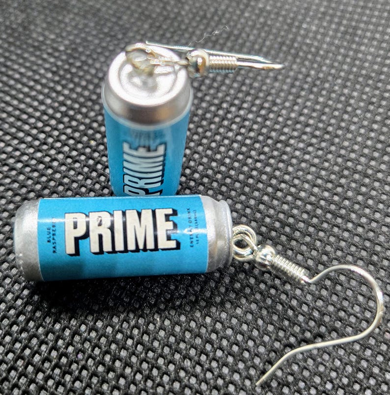 Puede incluir: Pendientes novedosos con forma de peque&ntilde;as latas de bebida energ&eacute;tica PRIME azules y plateadas. Los pendientes muestran la palabra "PRIME" en letras blancas sobre un fondo azul, con ganchos plateados.