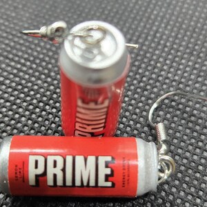 Puede incluir: Pendientes de lata de bebida energ&eacute;tica PRIME roja y plateada. Los pendientes presentan la palabra "PRIME" en blanco. Un pendiente est&aacute; tumbado y el otro est&aacute; de pie con un gancho plateado.  Texto "Lemon Lime".