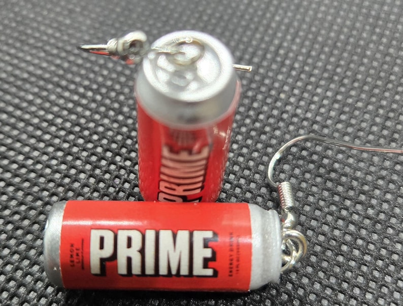 Puede incluir: Pendientes de lata de bebida energ&eacute;tica PRIME roja y plateada. Los pendientes presentan la palabra "PRIME" en blanco. Un pendiente est&aacute; tumbado y el otro est&aacute; de pie con un gancho plateado.  Texto "Lemon Lime".