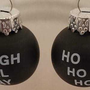 Può includere: Due palline di Natale nere con cime argentate. Una pallina dice "Sleigh All Day" e l'altra dice "Ho Ho Ho".