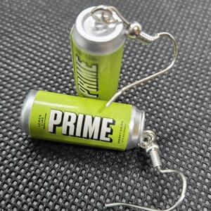 Puede incluir: Un par de pendientes originales con forma de peque&ntilde;as latas de bebida energ&eacute;tica PRIME de color verde lima y plata. Los pendientes muestran la palabra "PRIME" en blanco y las palabras "Lemon Lime" y "Energy Drink". Tienen ganchos plateados.