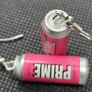 Puede incluir: Un par de pendientes novedosos con forma de peque&ntilde;as latas rosas y plateadas de bebida energ&eacute;tica PRIME. Los pendientes presentan la palabra "PRIME" en letras blancas y el sabor "Strawberry Watermelon". Los pendientes tienen ganchos plateados.