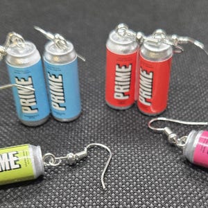 Puede incluir: Pendientes en miniatura con forma de latas de bebida PRIME en varios colores: verde, azul, rojo y rosa. Cada lata presenta la palabra "PRIME" en blanco. Los pendientes tienen cierres de gancho de pez plateados.