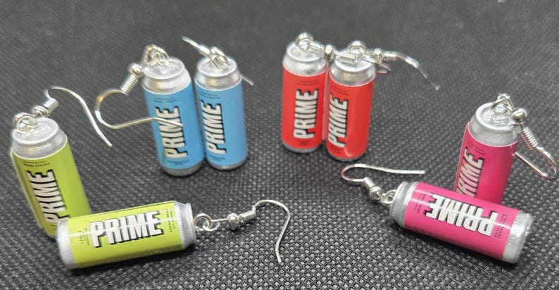 Puede incluir: Pendientes en miniatura con forma de latas de bebida PRIME en varios colores: verde, azul, rojo y rosa. Cada lata presenta la palabra "PRIME" en blanco. Los pendientes tienen cierres de gancho de pez plateados.