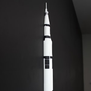 May include: A white and black model of a Saturn V rocket, a symbol of space exploration. The model is vertically oriented against a dark background, showcasing its detailed design. The rocket's height is approximately 36 inches.