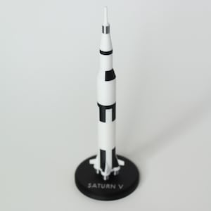 May include: A model of the Saturn V rocket, predominantly white with black accents and a silver tip. The rocket stands on a black circular base with the text "SATURN V". This collectible item is a detailed replica of the historic spacecraft.