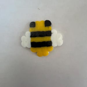 Pode incluir: Criação de contas em forma de abelha amarela, preta e branca. A abelha tem um corpo amarelo com listras pretas, detalhes da cabeça pretos e asas brancas. O item é provavelmente feito de contas de plástico.